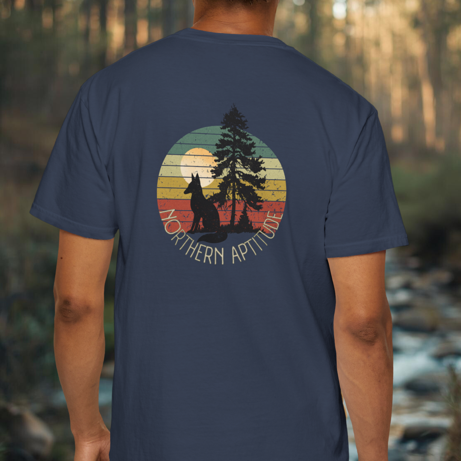 Person wearing a midnight blue t-shirt with a graphic design of a fox, pine and sunset on the back, set against a forest background.