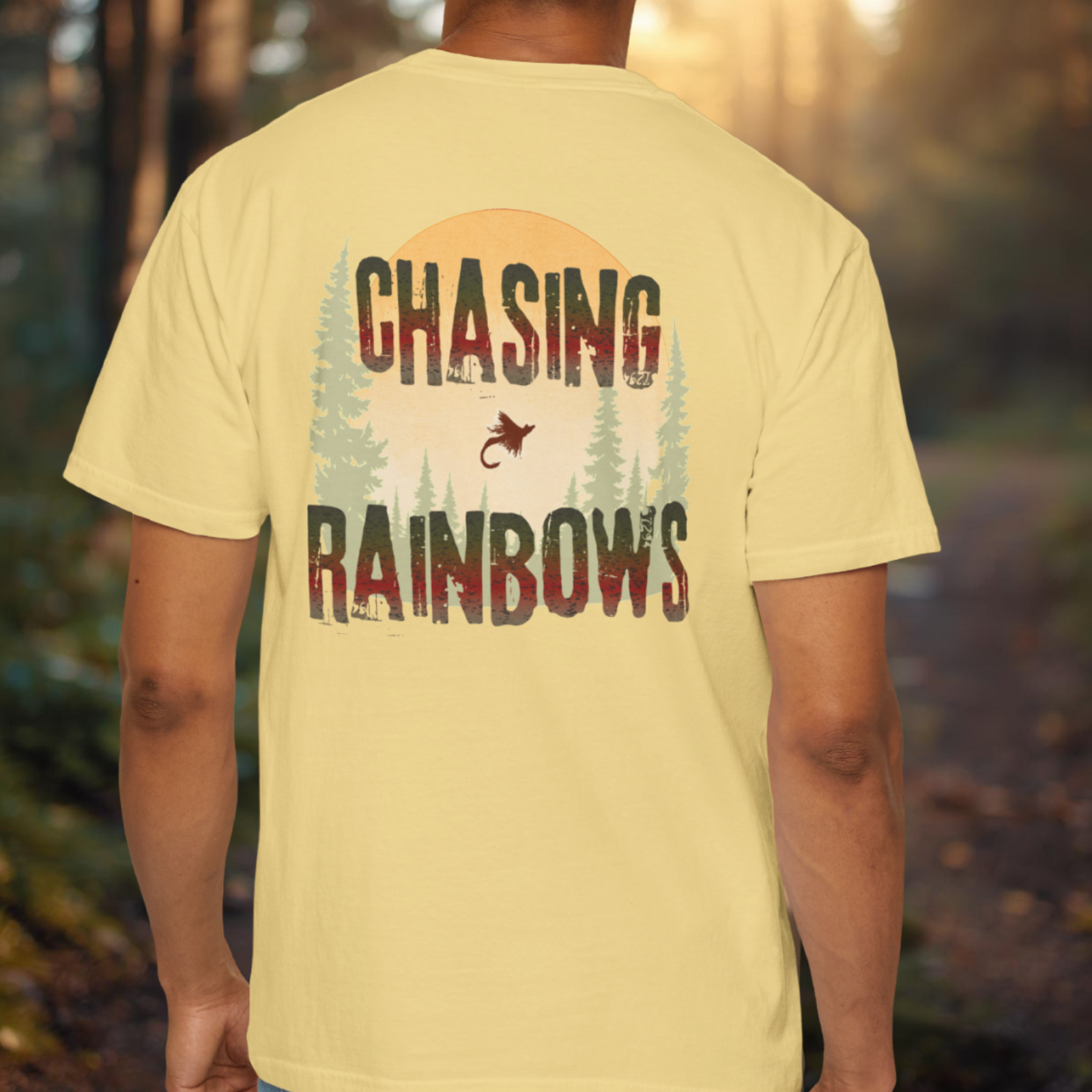Person wearing a butter colored t-shirt with 'Chasing Rainbows' text and graphic in a forest setting