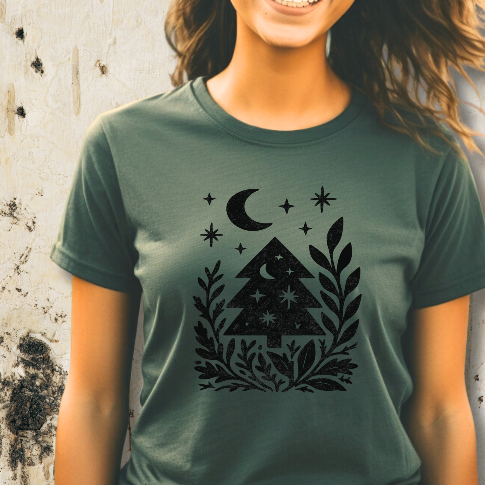 Woman wearing blue spruce forest graphic t-shirt with charcoal grey trees and crescent moon, standing against rustic wall.