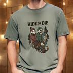 Person wearing moss green “Ride or Die” skeleton sledding graphic t-shirt in front of wood wall with string lights