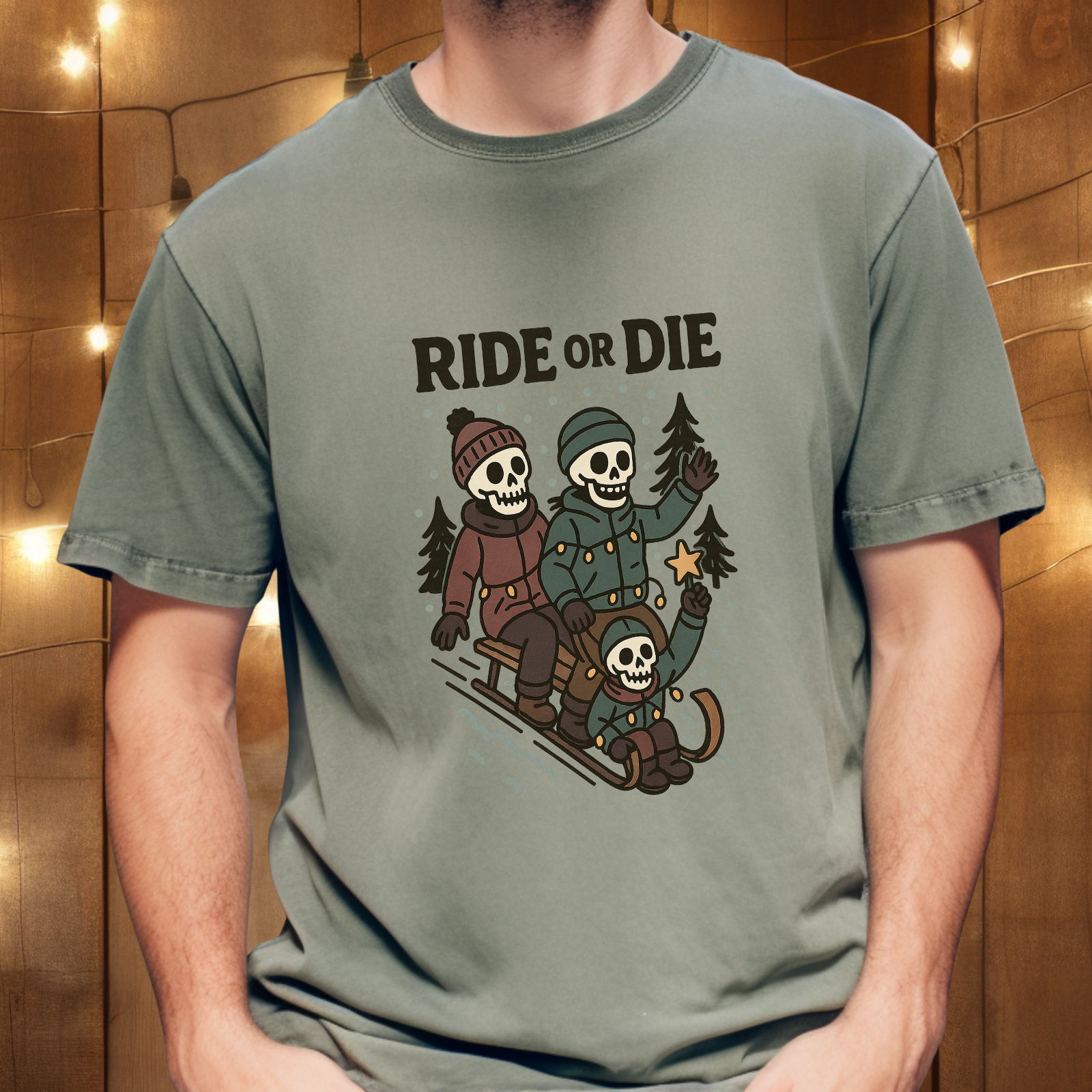 Person wearing moss green “Ride or Die” skeleton sledding graphic t-shirt in front of wood wall with string lights
