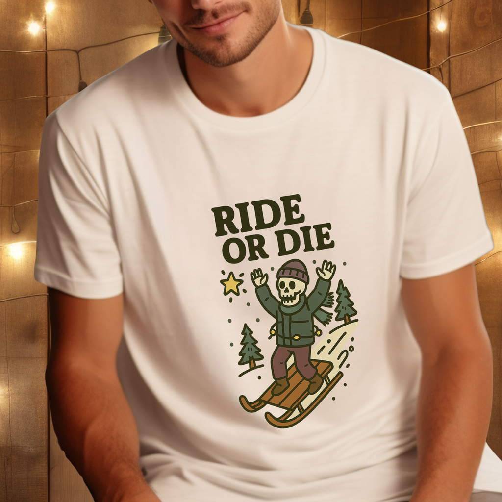 Person wearing white “Ride or Die” skeleton sledding graphic t-shirt in a cozy setting with warm string lights in the background.