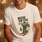 Person wearing white “Ride or Die” skeleton sledding graphic t-shirt in a cozy setting with warm string lights in the background.