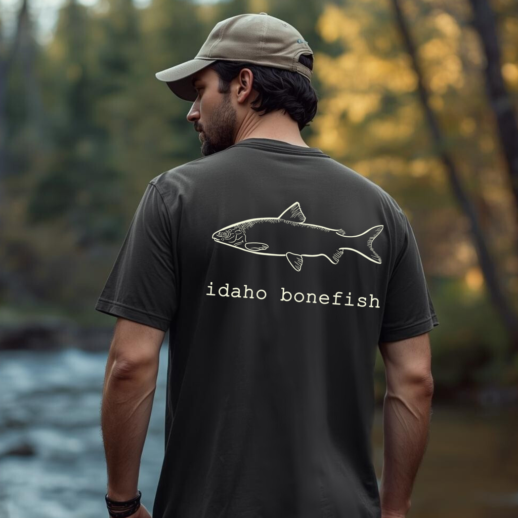 Fly fisher wearing 'Idaho Bonefish' hand drawn river whitefish sketch graphic t-shirt near water.
