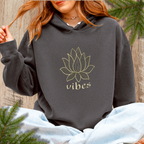 Person wearing a dark pepper hoodie with a lotus flower design and 'vibes' text, sitting against a wooden background with greenery.