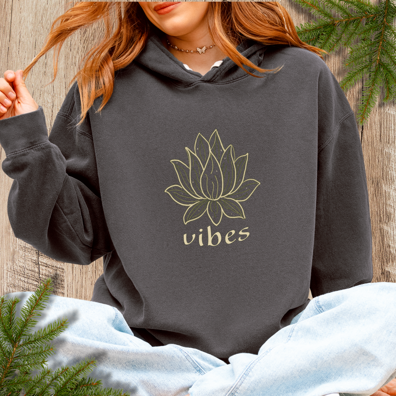 Person wearing a dark pepper hoodie with a lotus flower design and 'vibes' text, sitting against a wooden background with greenery.