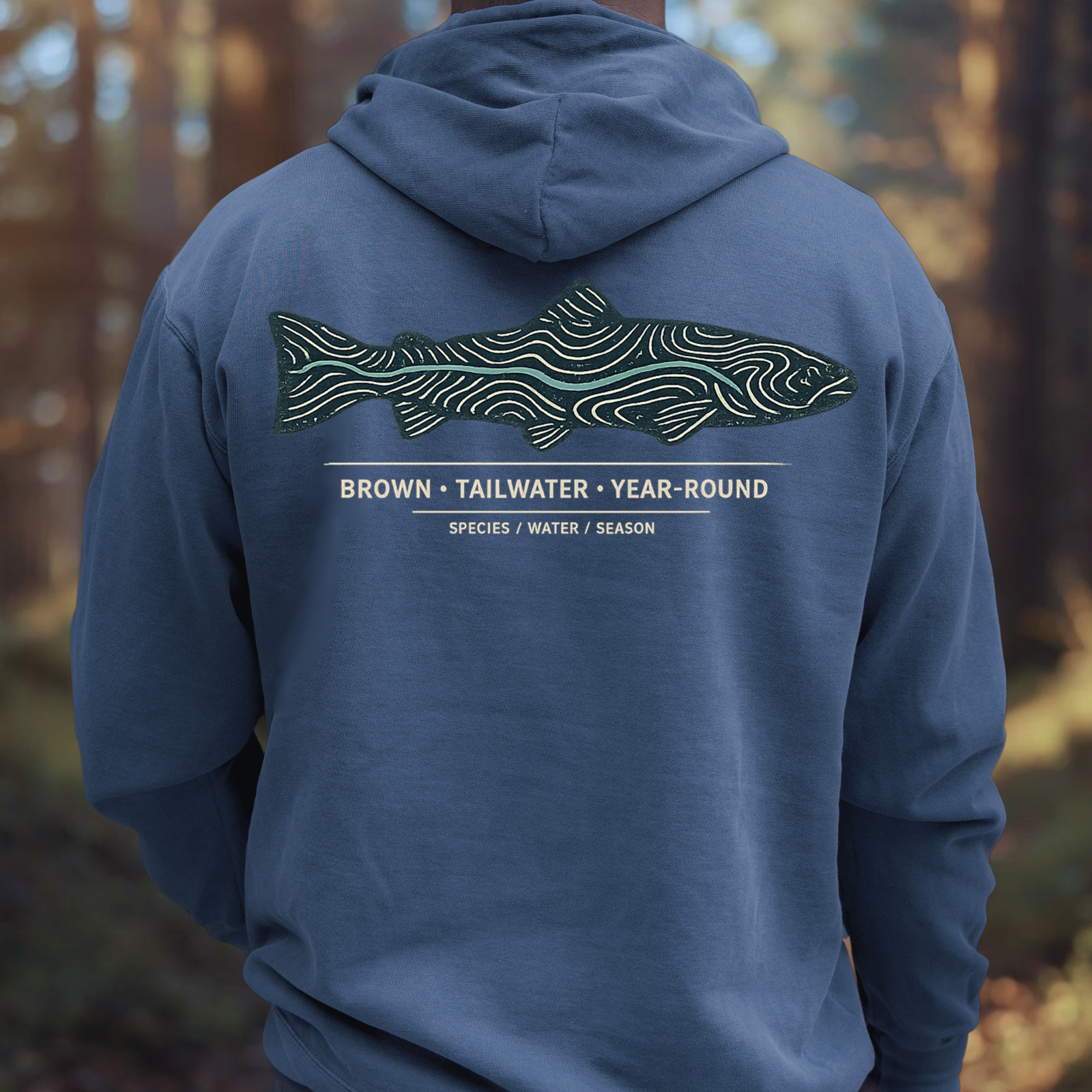 Person wearing brown trout topography hoodie, showing back trout graphic while standing outdoors by the water.