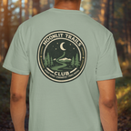 Person wearing a green t-shirt with 'Moonlit Trails Club' design in a forest setting