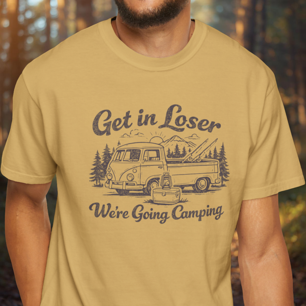 Person wearing a mustard yellow t-shirt with a graphic and text 'Get in Loser, We're Going Camping' in a forest setting.