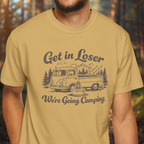 Person wearing a mustard yellow t-shirt with a graphic and text 'Get in Loser, We're Going Camping' in a forest setting.
