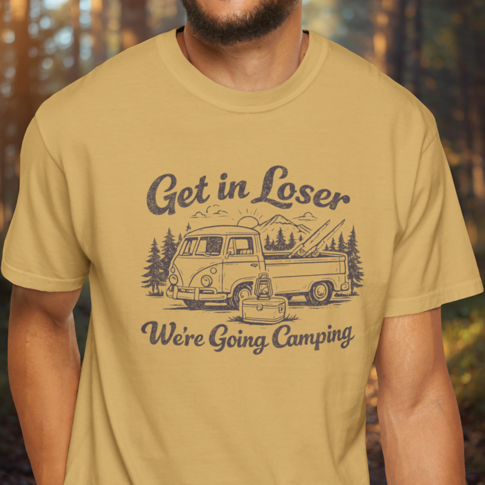 Person wearing a mustard yellow t-shirt with a graphic and text 'Get in Loser, We're Going Camping' in a forest setting.