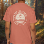 Person wearing a terra cotta t-shirt with a back print River Access Pass circular fishing emblem old map style in a forest setting