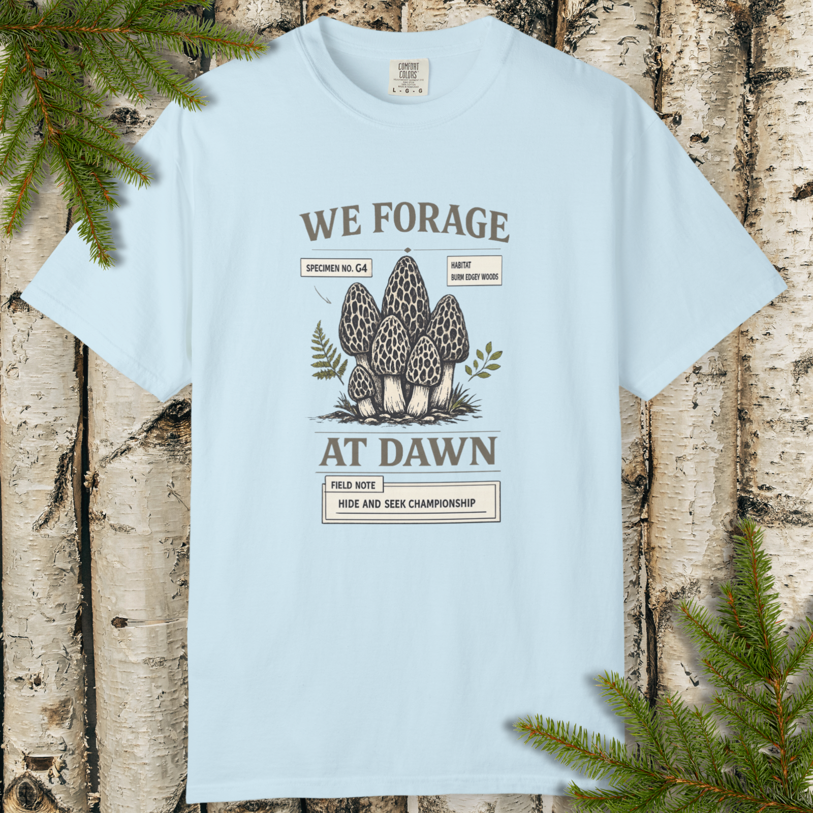 We Forage at Dawn morel mushroom graphic t-shirt flat lay on wood background.