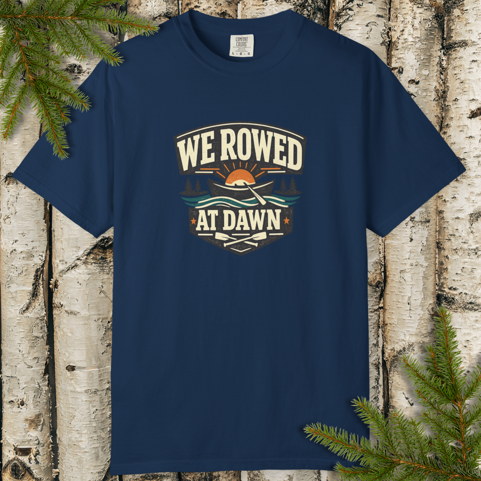 Navy blue t-shirt with 'We Rowed at Dawn' graphic on a wooden background with pine branches.