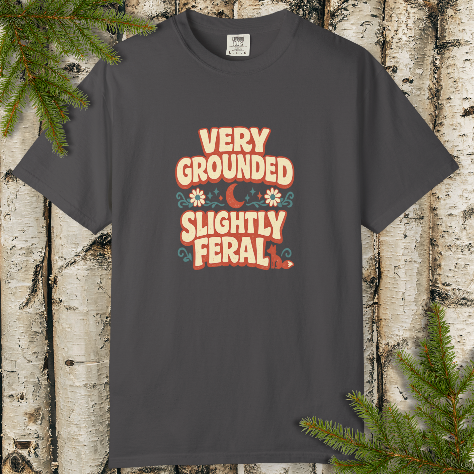 Gray t-shirt with 'Very Grounded Slightly Feral' text on a wooden background with pine branches.