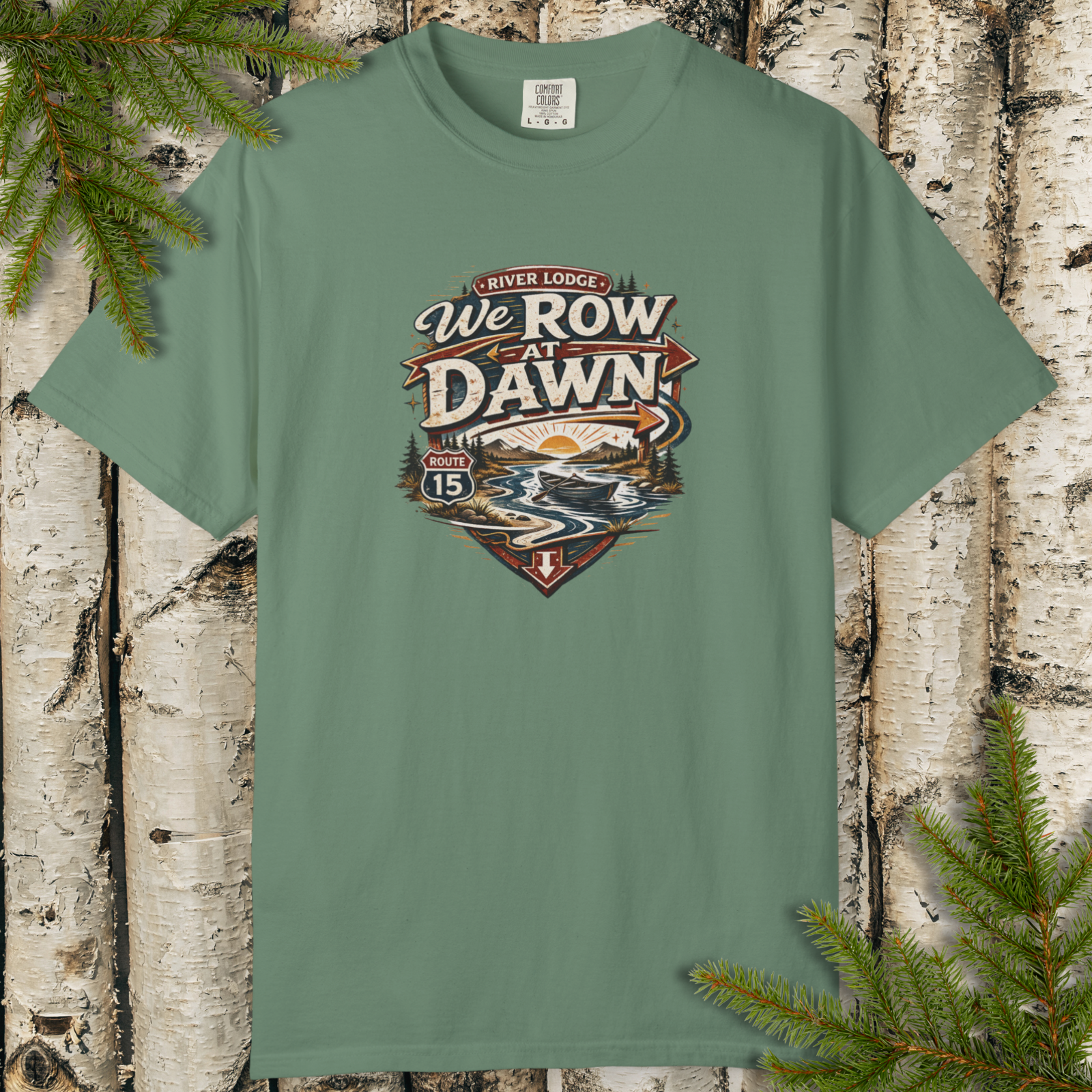 Green t-shirt with 'We Row at Dawn' design on a wooden background with pine branches.