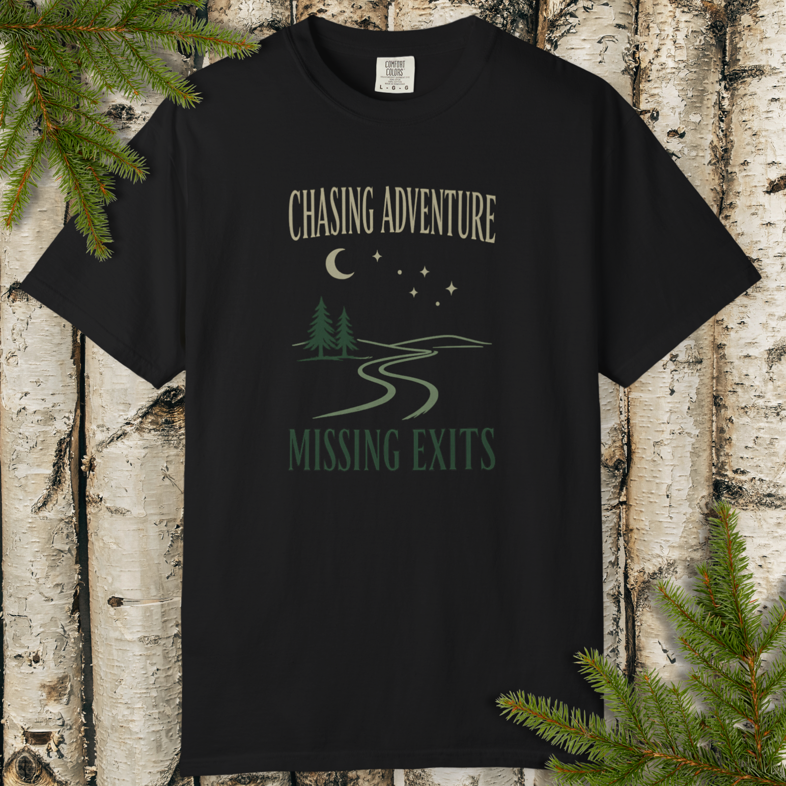 Black t-shirt with 'Chasing Adventure Missing Exits' design on a wooden background with pine branches.