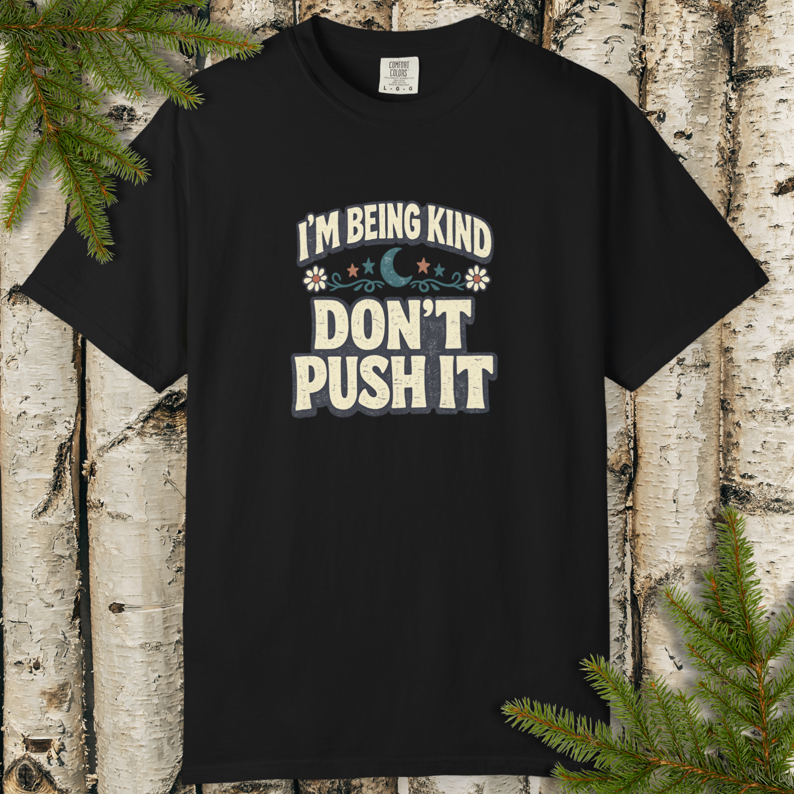 Black t-shirt with 'I'm being kind, don't push it' text on a wooden background with greenery.