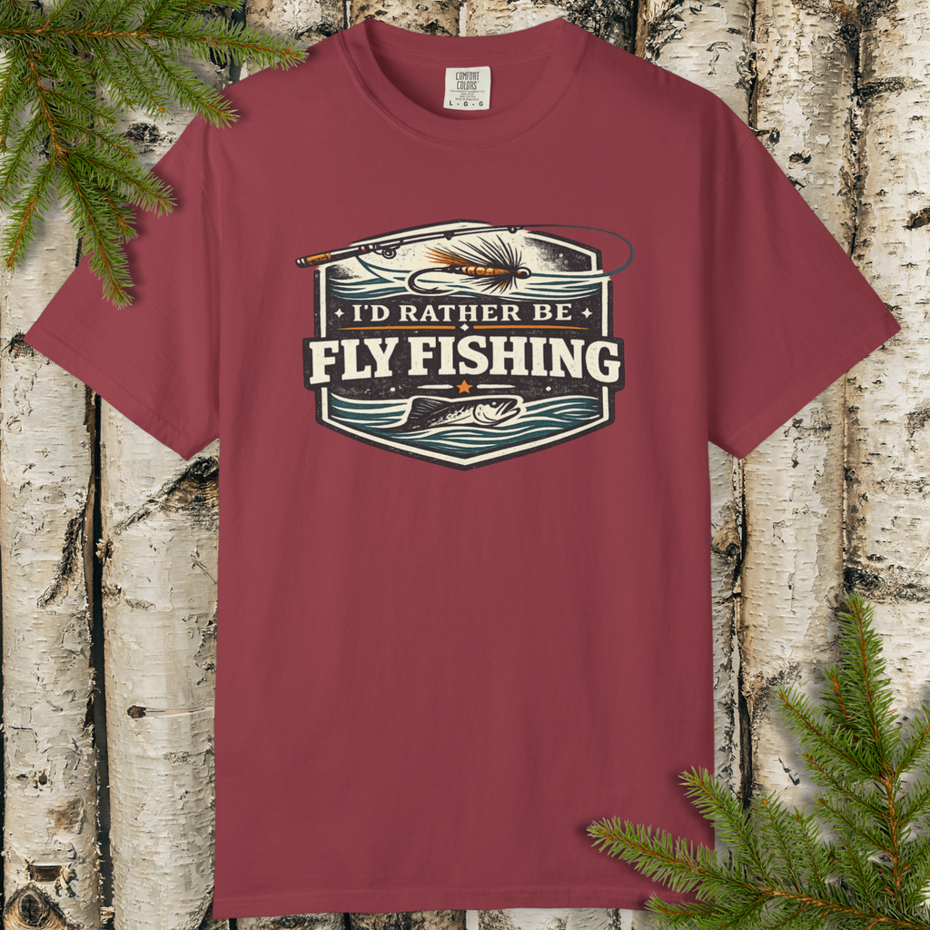 Maroon t-shirt with 'I'd Rather Be Fly Fishing' graphic on a wooden background with pine branches.
