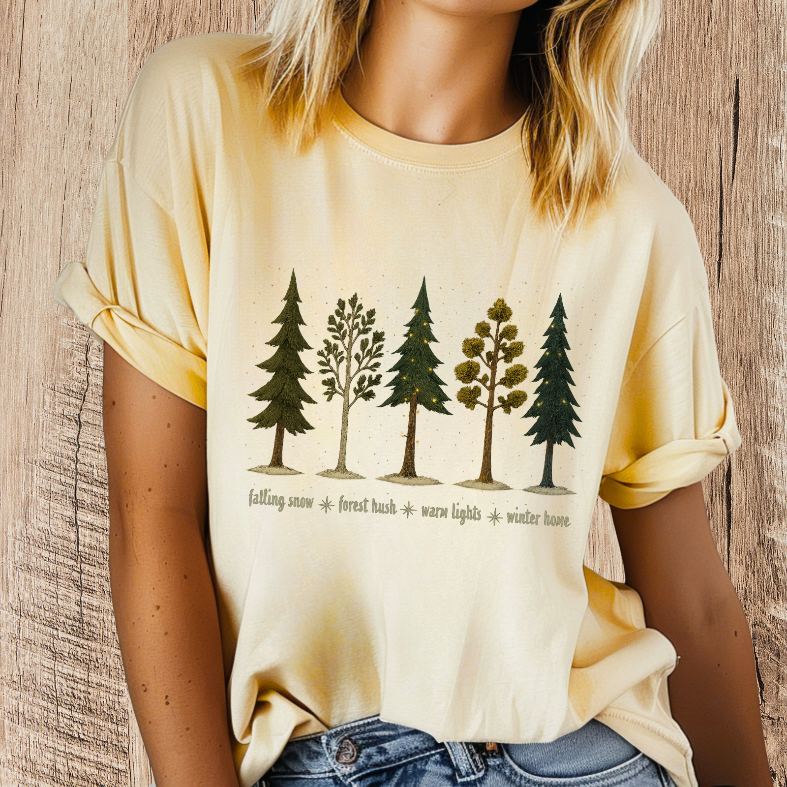 “Woman wearing pale yellow t-shirt with row of illustrated evergreen trees and ‘falling snow • forest hush • warm lights • winter home’ text.”