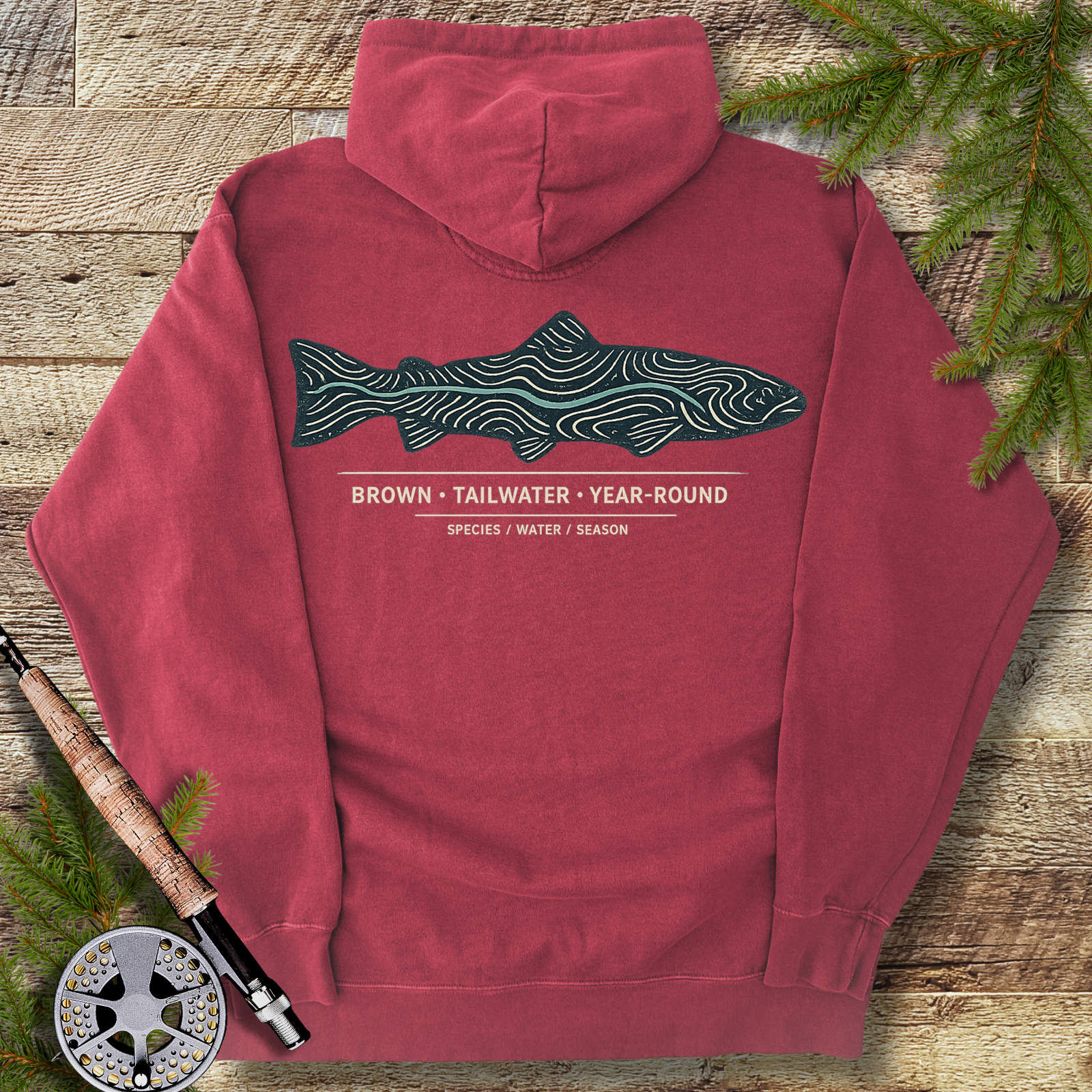 Crimson red hoodie with brown trout topography line-art graphic on back, styled on wooden background with fly-fishing gear.