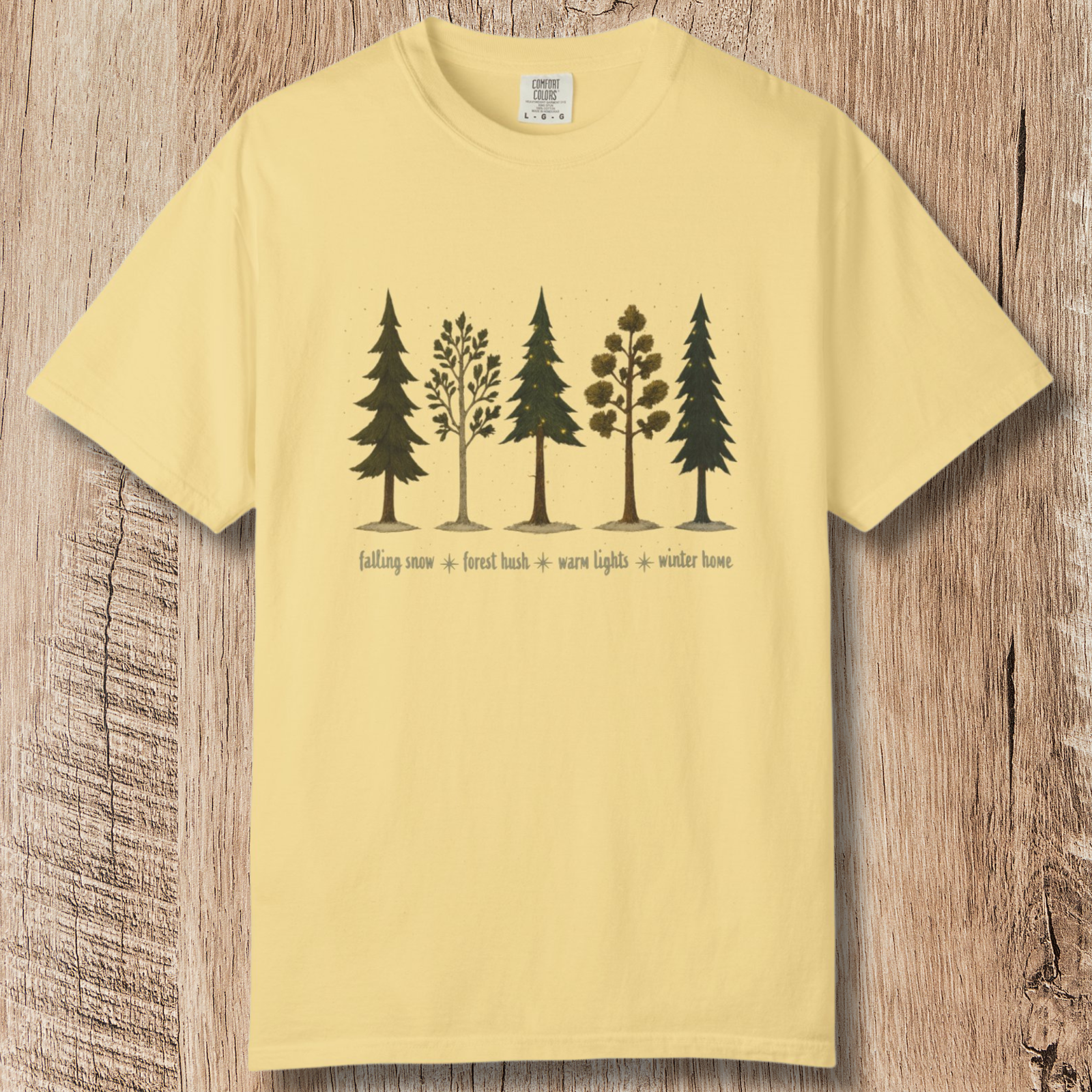 Yellow tee with illustration of five trees with different styles on a beige background, featuring text 'falling snow', 'forest hush', 'warm lights', and 'winter home'.
