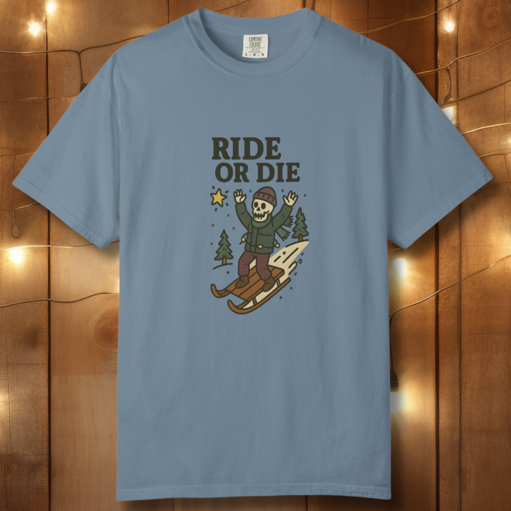 Ice blue t-shirt with “Ride or Die” text and skeleton sledding graphic, displayed against wood backdrop with string lights.