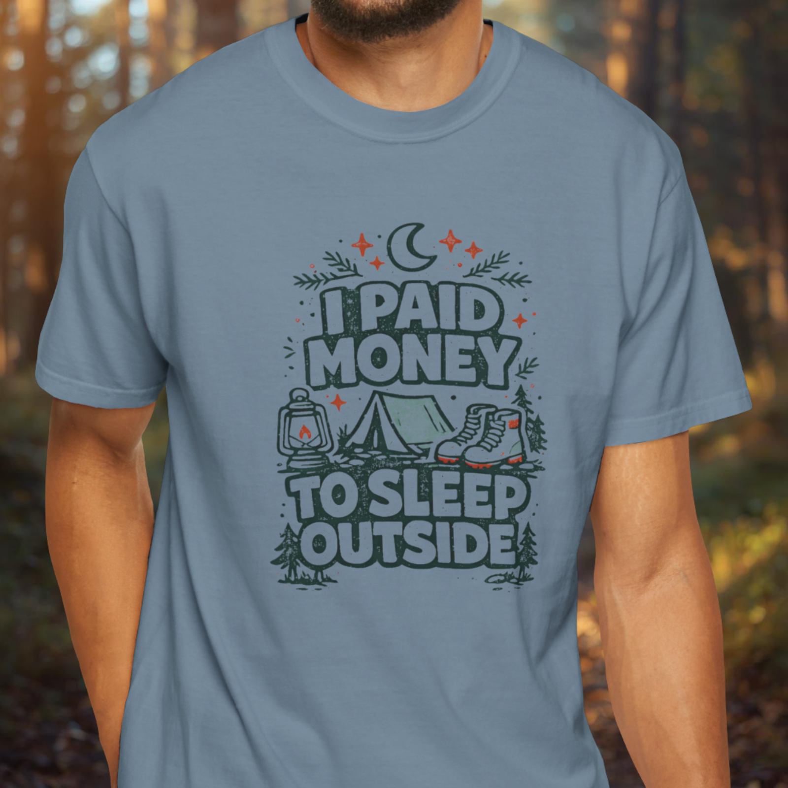 Person wearing a blue t-shirt with a camping-themed graphic and text reading “I Paid Money to Sleep Outside” with rustic outdoor design.