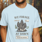 Light blue t-shirt with mushroom graphic and text 'We Forage at Dawn' worn by a person in a forest setting.