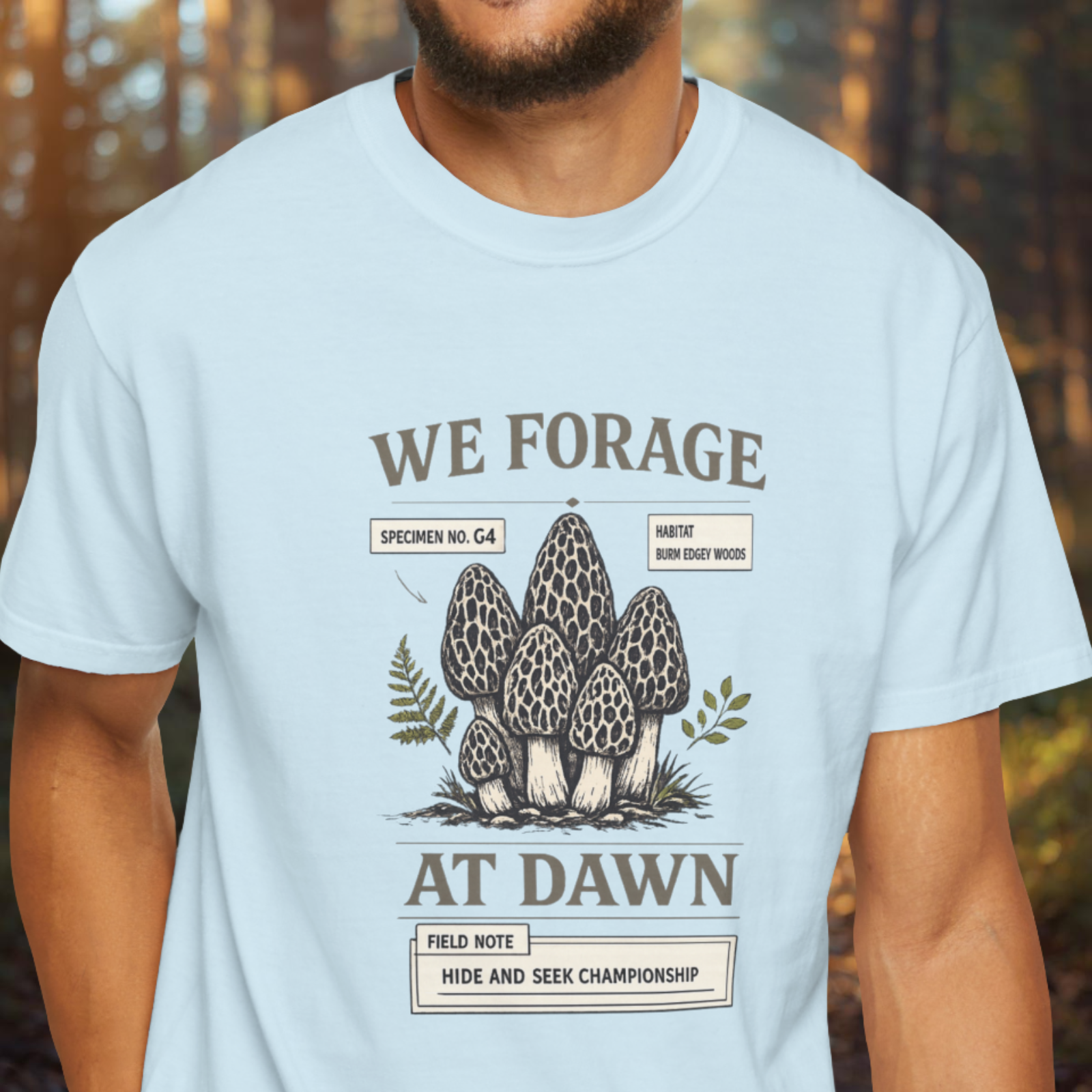 Light blue t-shirt with mushroom graphic and text 'We Forage at Dawn' worn by a person in a forest setting.