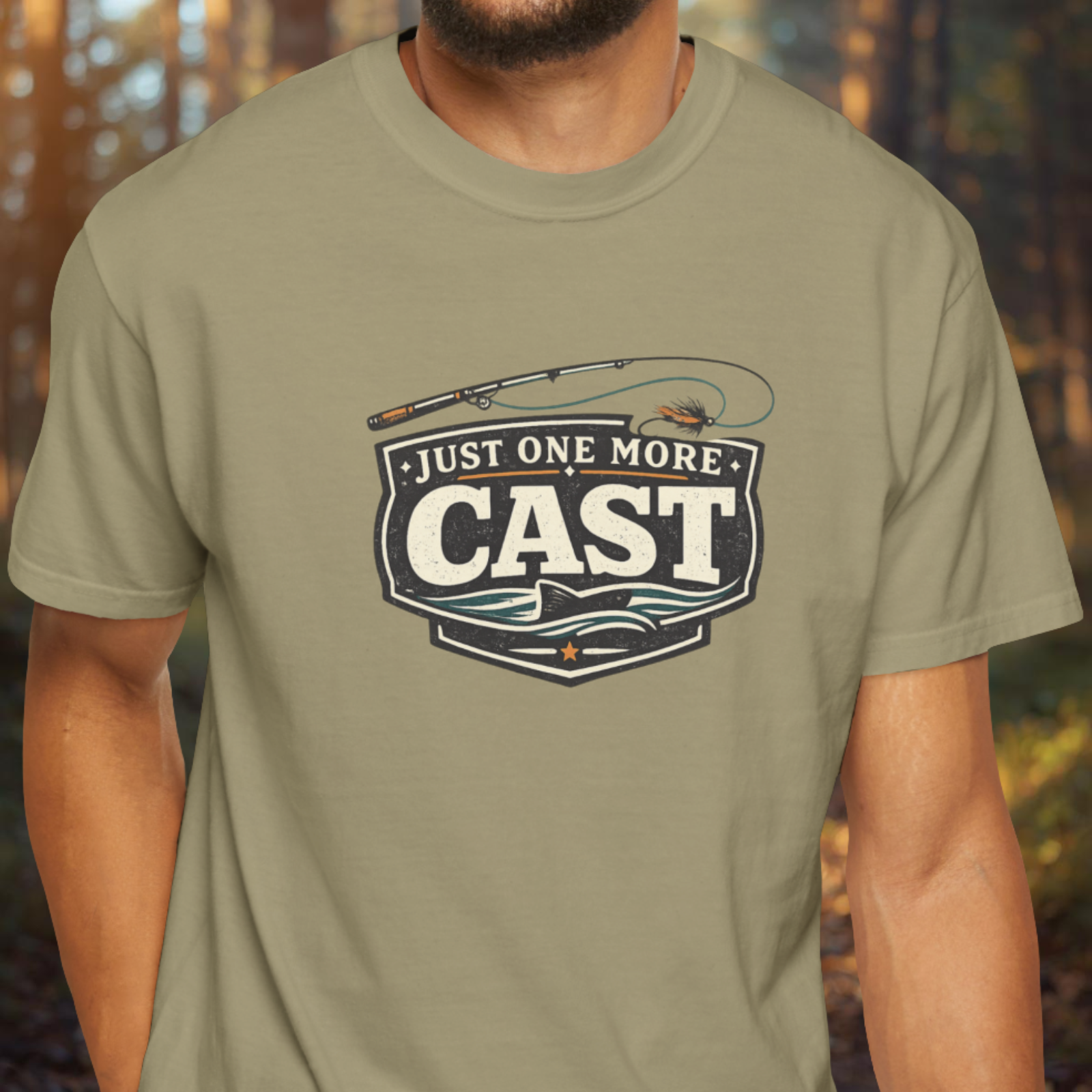 Person wearing a khaki t-shirt with a vintage “Just One More Cast” fishing badge graphic t-shirt in a relaxed outdoor angler style.