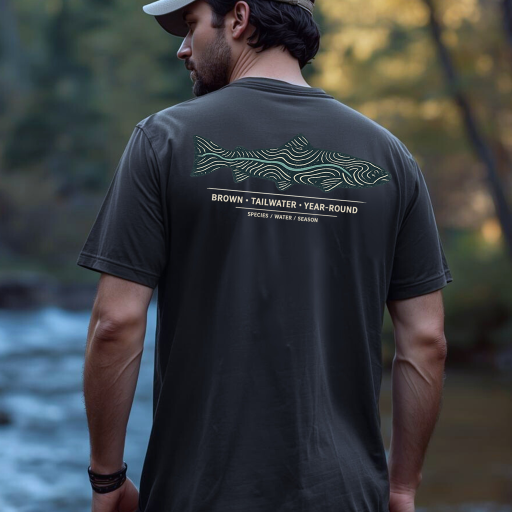 Person wearing a brown trout topography graphic t-shirt outdoors near the water.