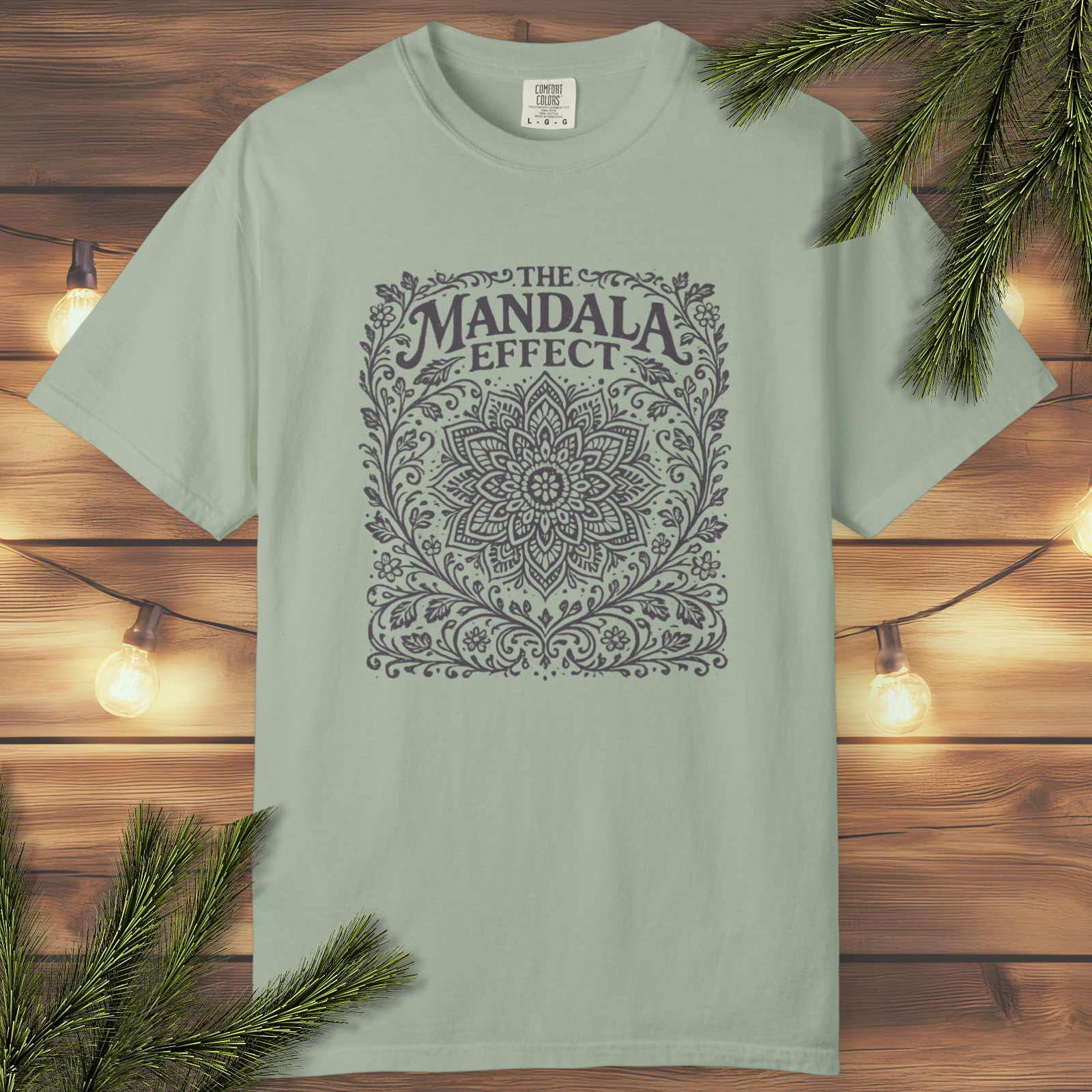 Light green t-shirt with 'The Mandala Effect' design on a wooden background with lights and branches.