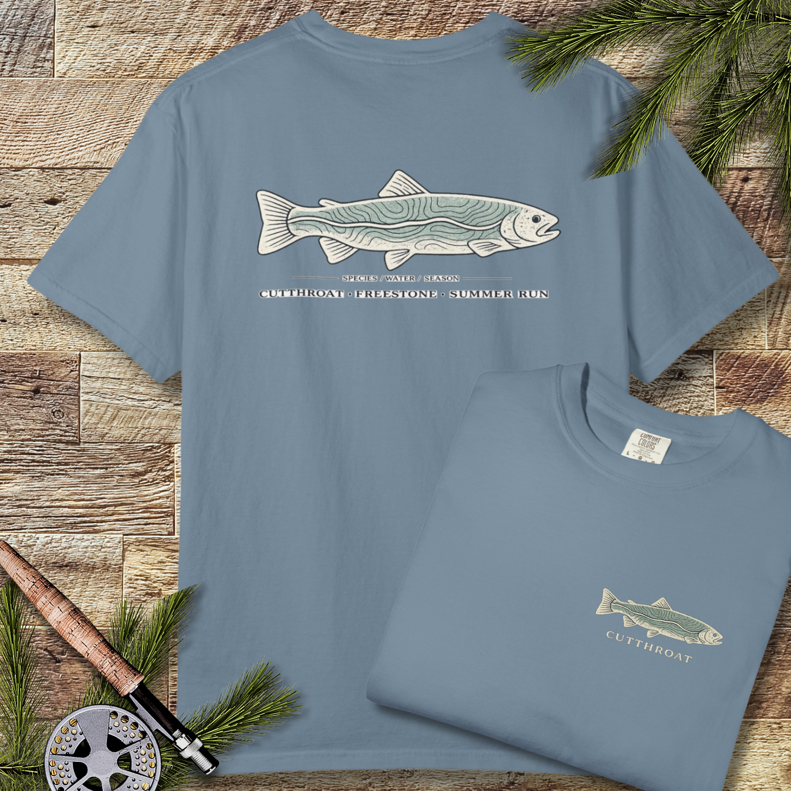 Garment-dyed ice blue t-shirt with large cutthroat trout topography graphic across the back, laid flat on a wood background