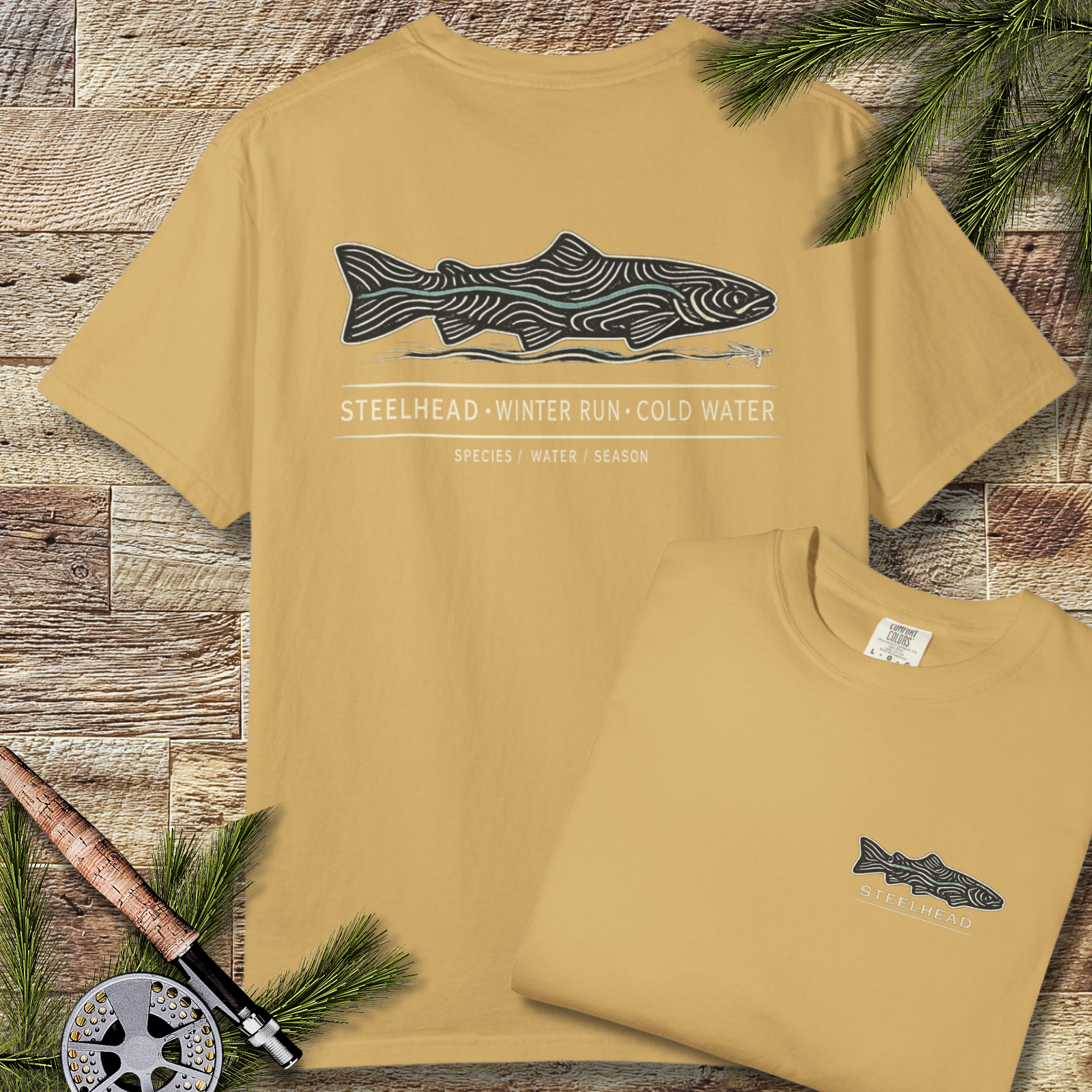 Mustard garment-dyed steelhead fishing t-shirt with small chest fish and large winter-run graphic on back, laid flat on wood with pine branches.