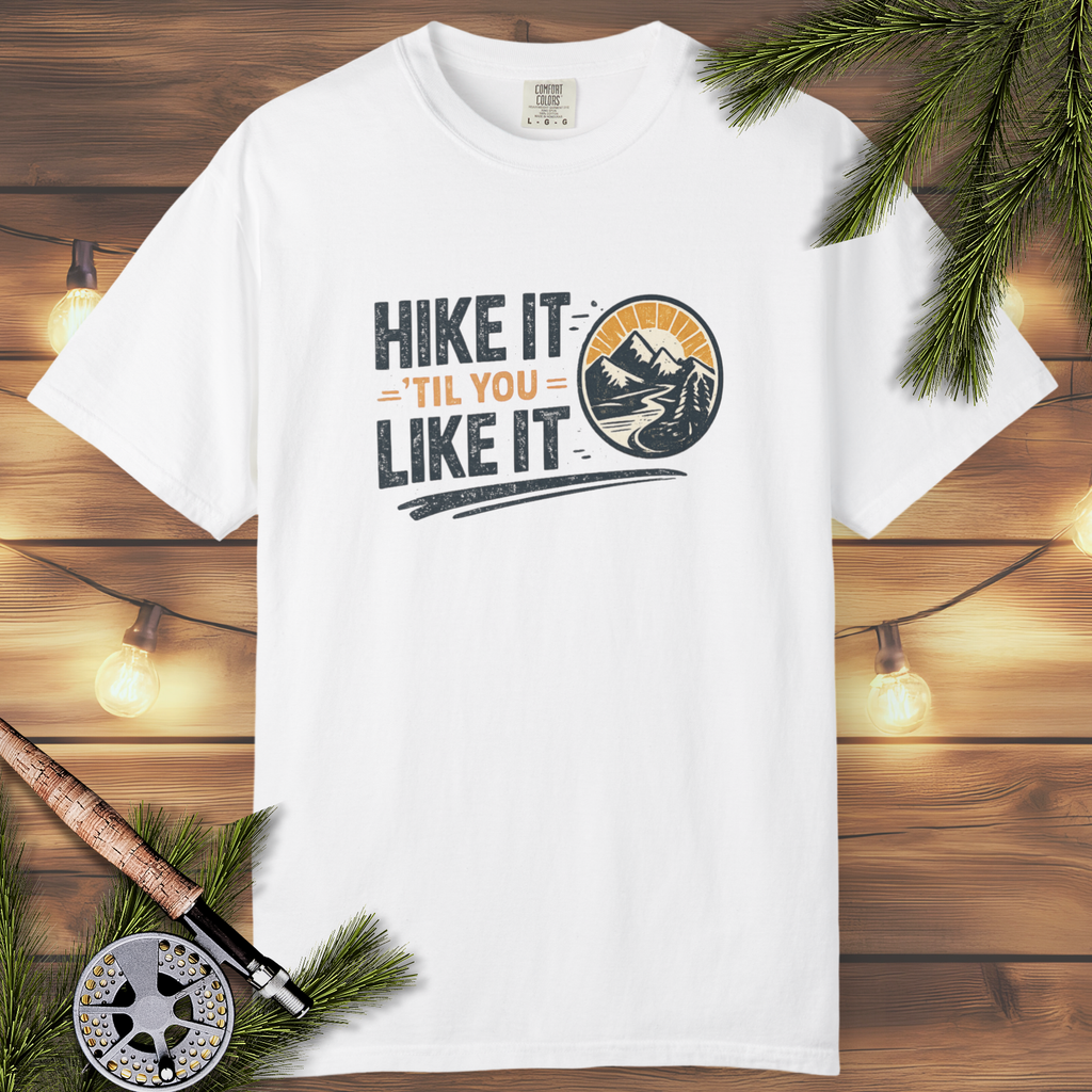 White t-shirt with 'Hike It 'Til You Like It' design on a wooden background with fishing equipment.