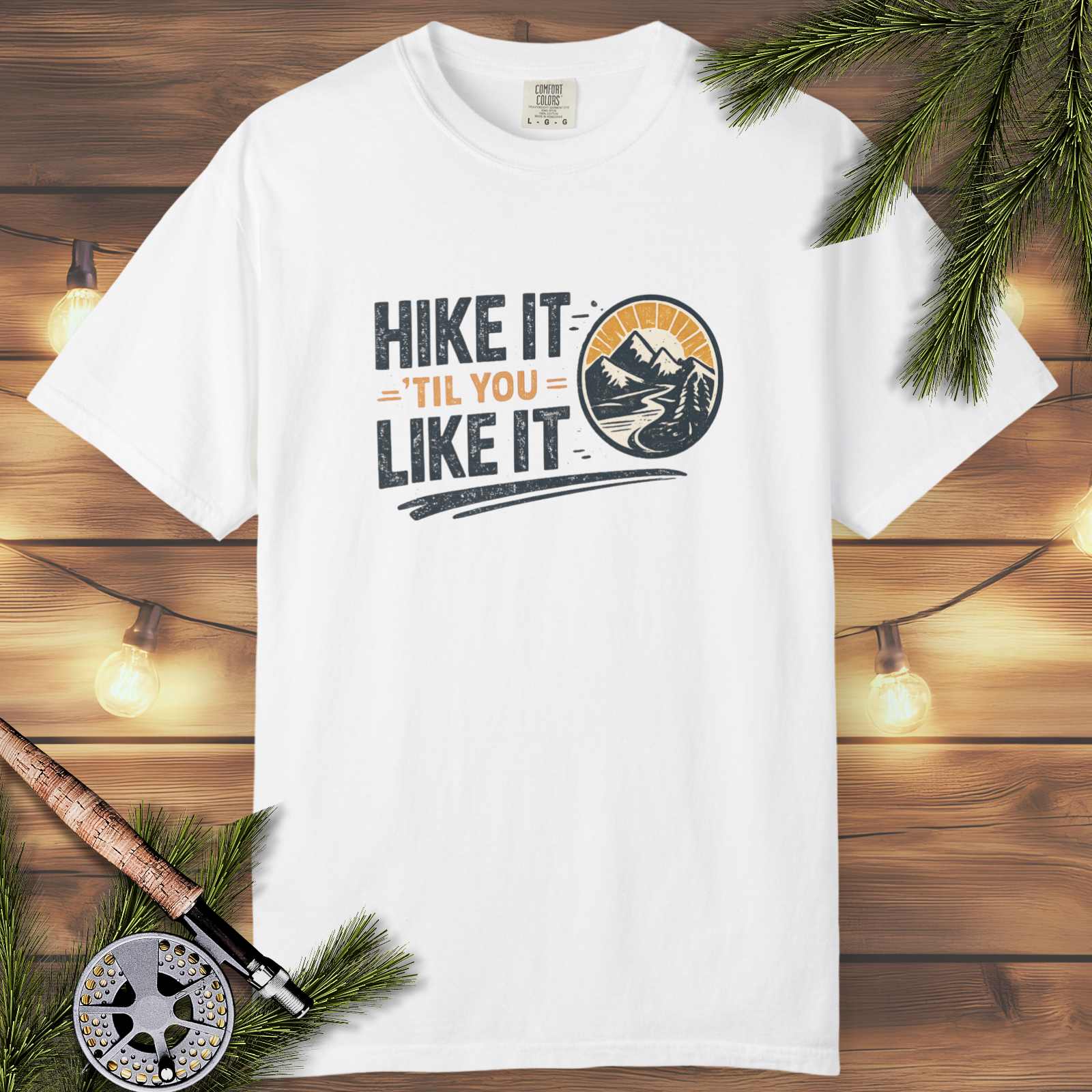 White t-shirt with 'Hike It 'Til You Like It' design on a wooden background with fishing equipment.