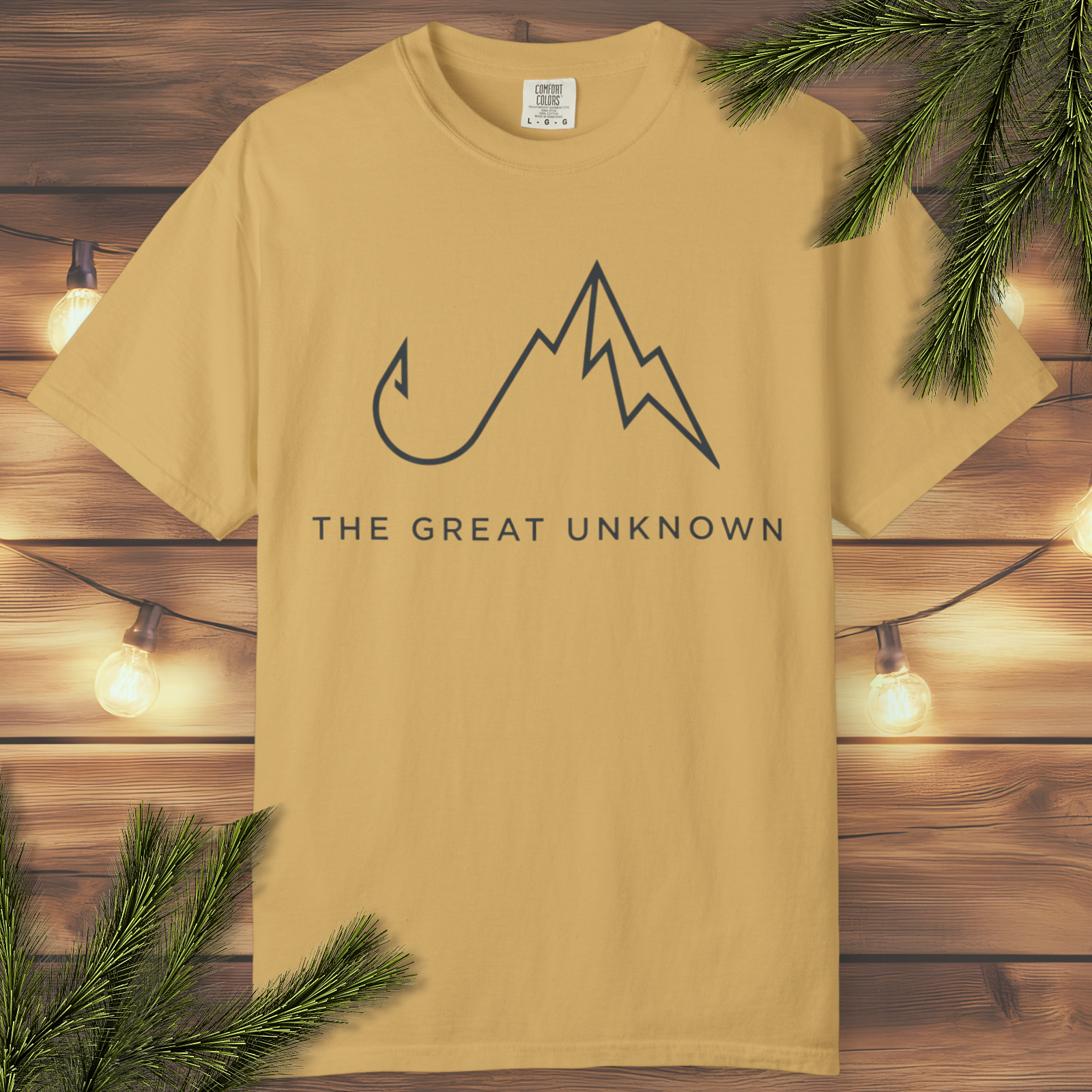 Mustard yellow t-shirt with mountain and hook design and 'The Great Unknown' text on a wooden background.