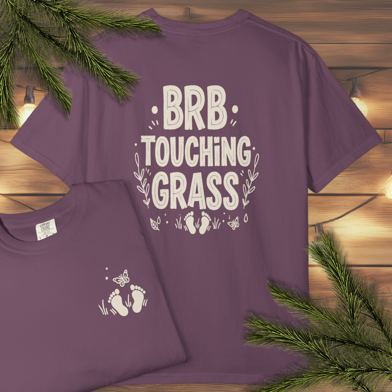 Berry t-shirt with back print 'BRB Touching Grass' hand-lettered foliage graphic on a wooden surface with greenery. 