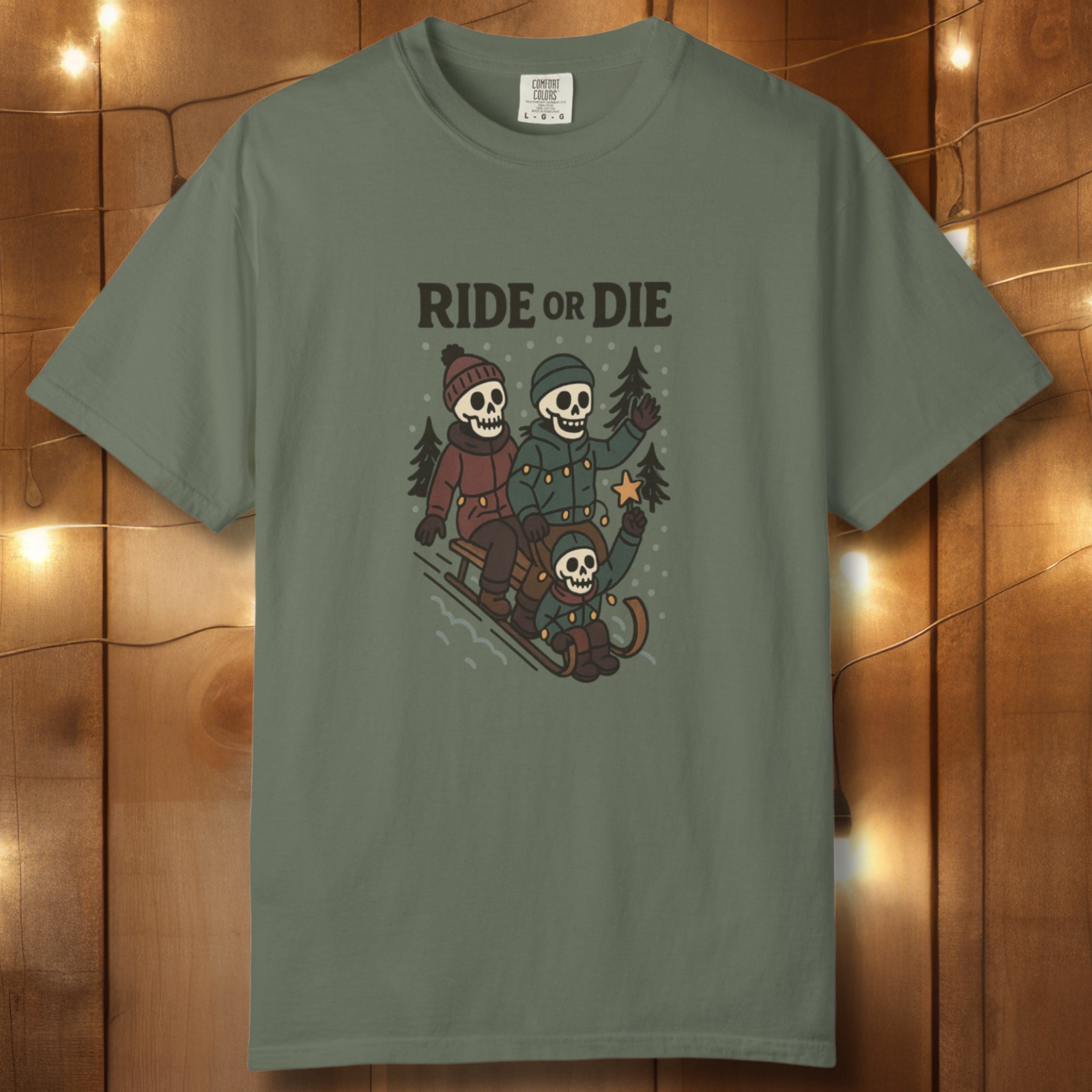 Moss green unisex t-shirt with “Ride or Die” skeleton sledding graphic on a wood backdrop with string lights