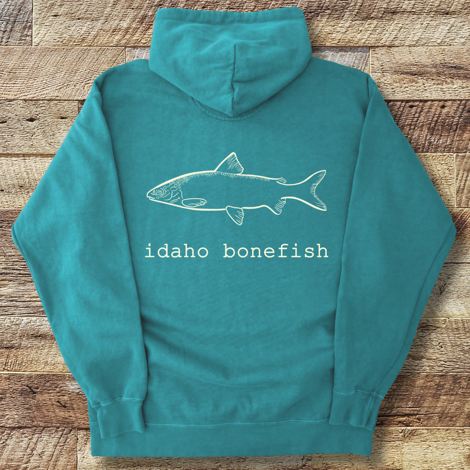 Seafoam hoodie with 'idaho bonefish' design on the back, placed on a wooden surface.